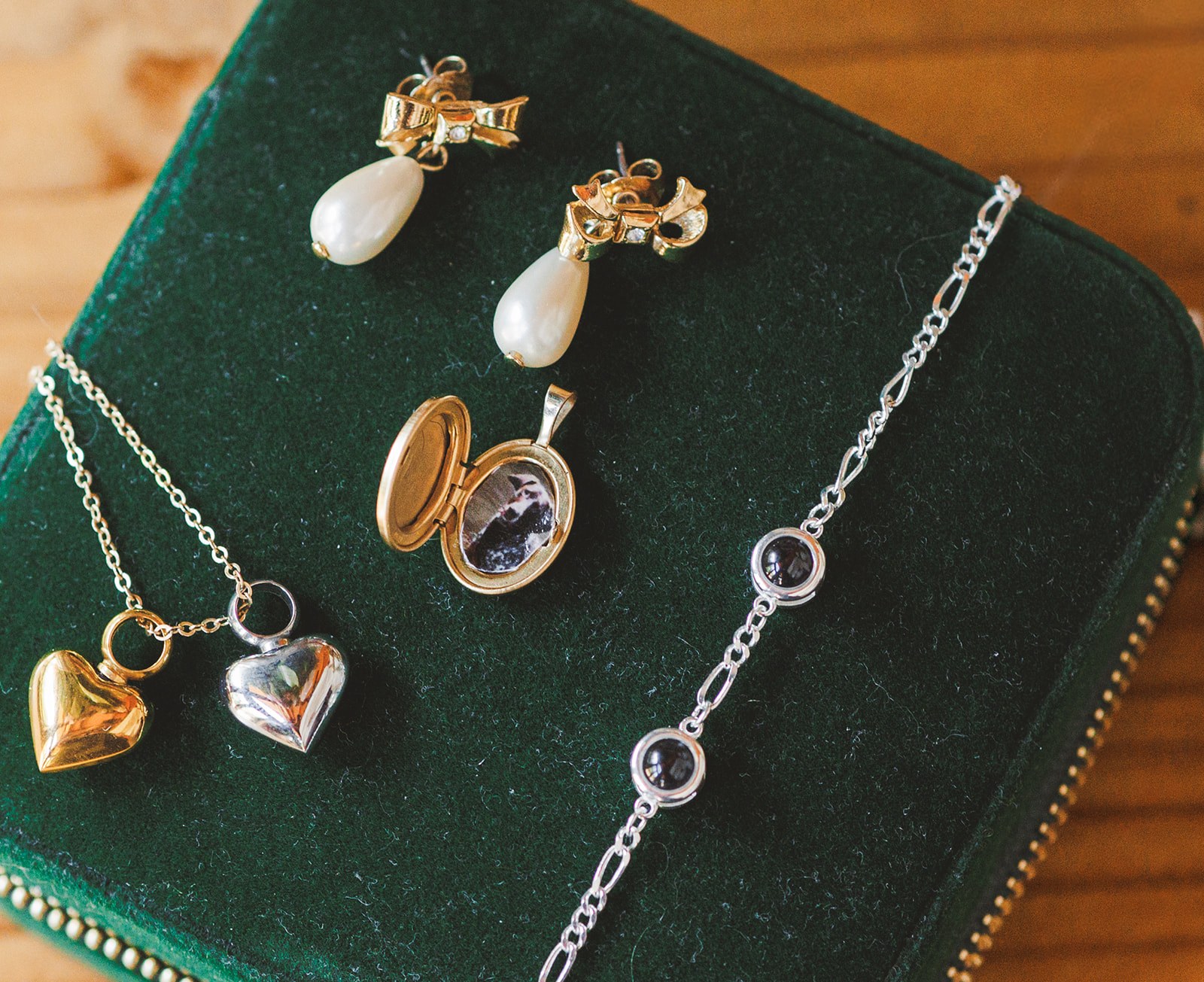 Pearl drop earrings, a small gold heart pendant, a silver heart, and an open locket with photographs inside, laid out on a green velvet jewelry case