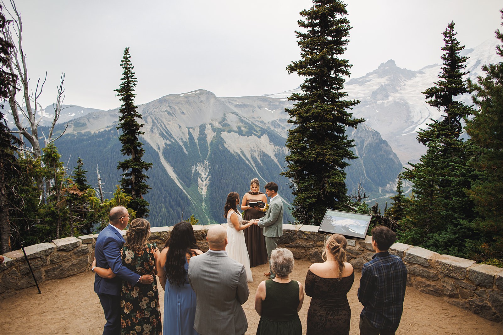 Michaela and AJ joining hands in front of their officiant on the stone terrace at Emmons Vista, their guests standing in a half circle with their backs to the camera and the glaciers of Mount Rainier filling the view behind