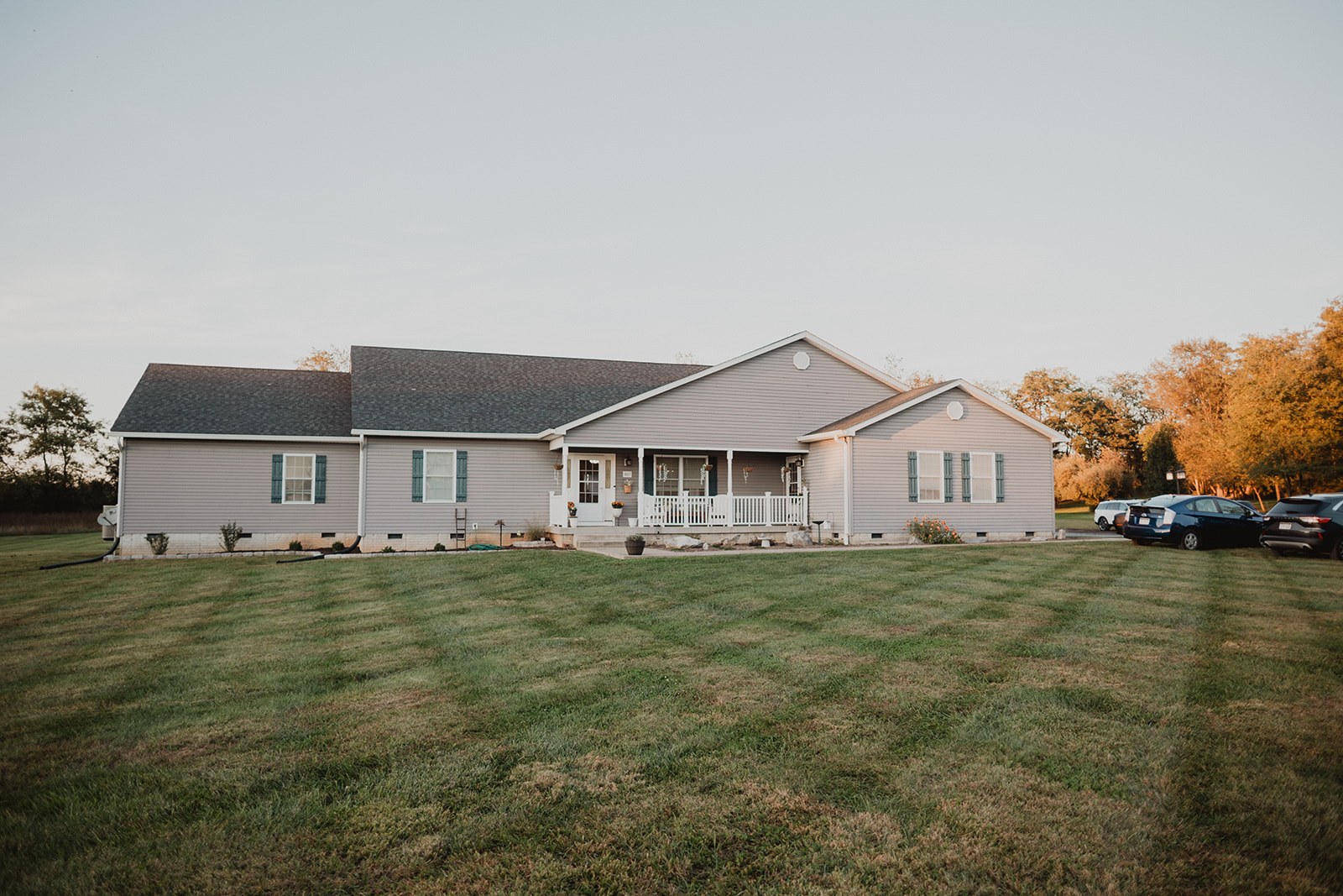 The house seen from the far side of the mown back lawn at golden hour, cars parked along the drive and treeline behind