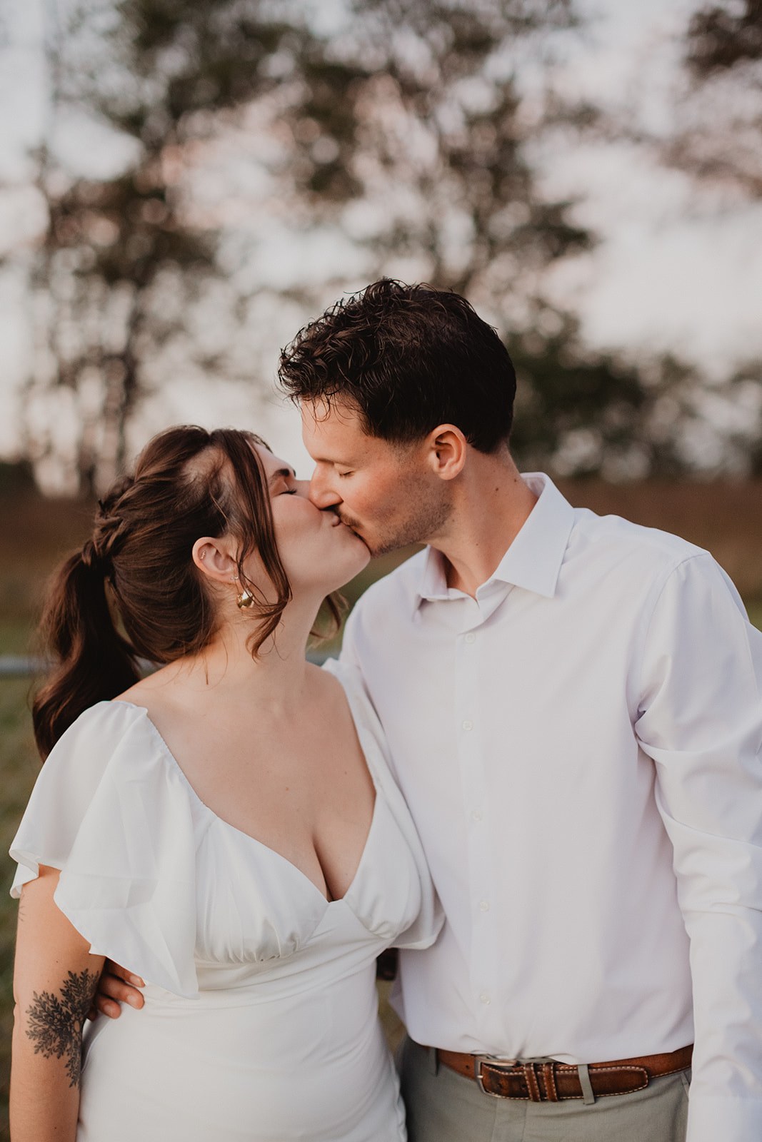 Michaela and AJ kissing on the lawn at dusk, bare trees and the last pale light behind them