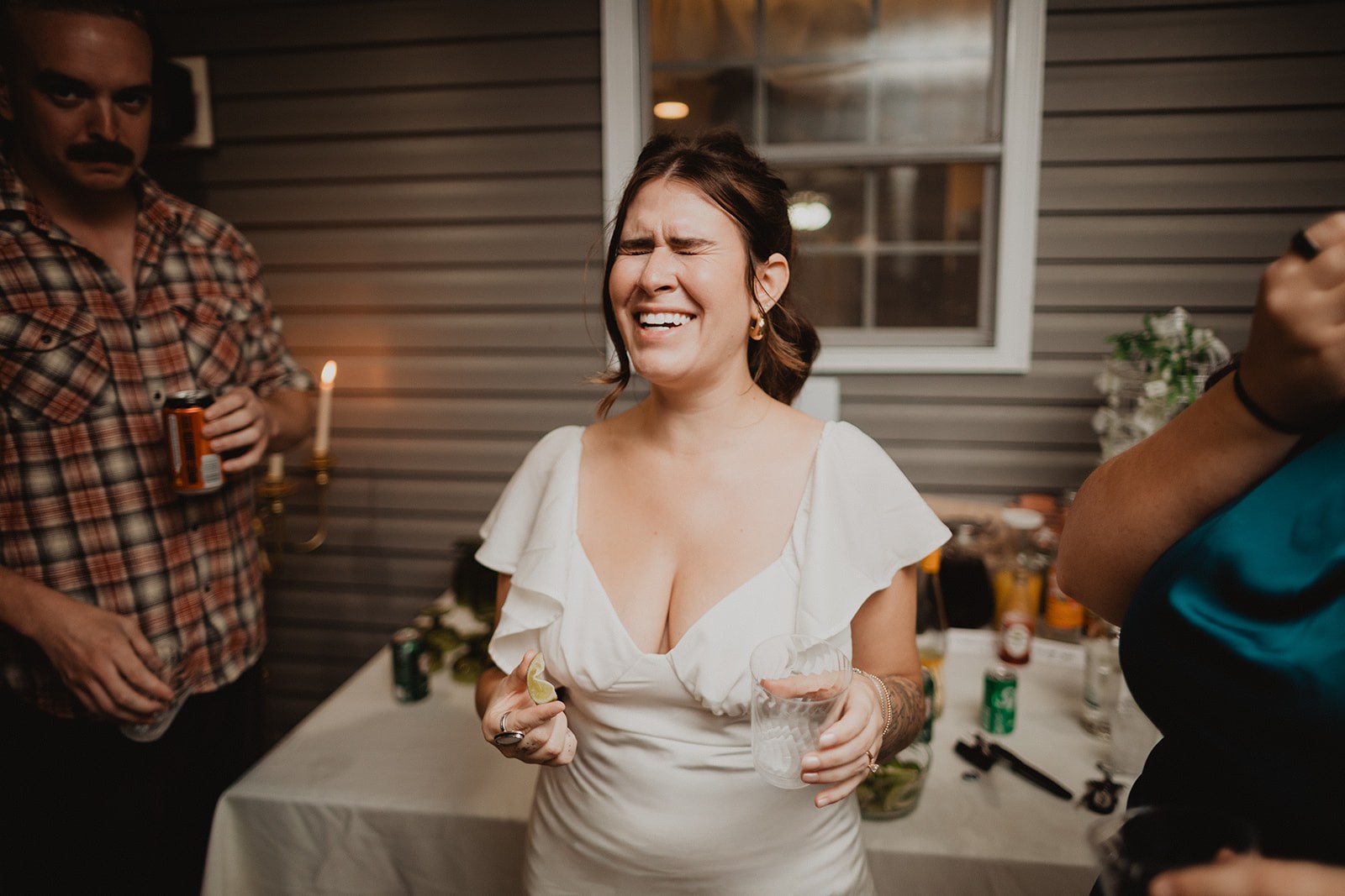 Michaela laughing hard mid sentence at the drinks table with a guest beside her and candles lit behind