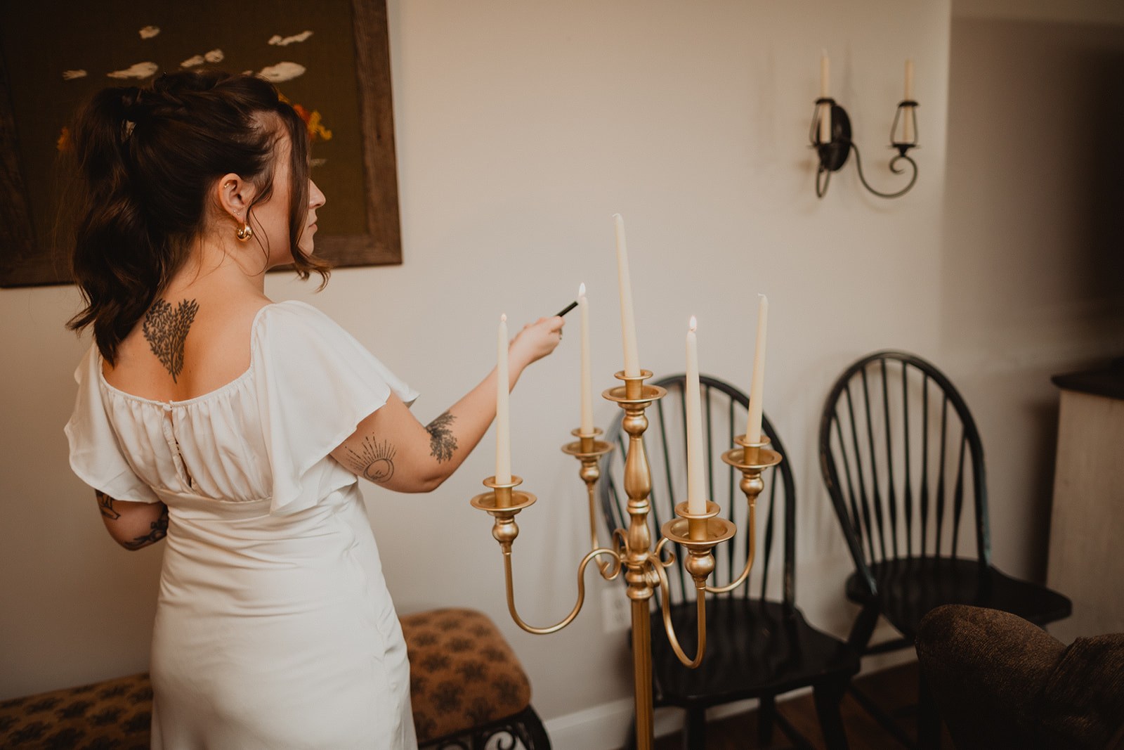 Michaela in her short white reception dress reaching up to light the tapers in a brass candelabra indoors before guests arrive