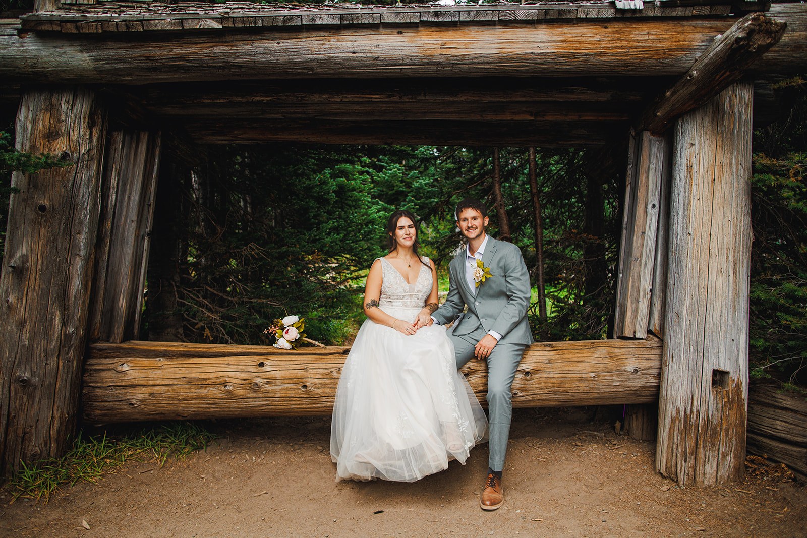 Michaela and AJ sitting side by side under the weathered log frame of an old trail shelter, her bouquet in her lap