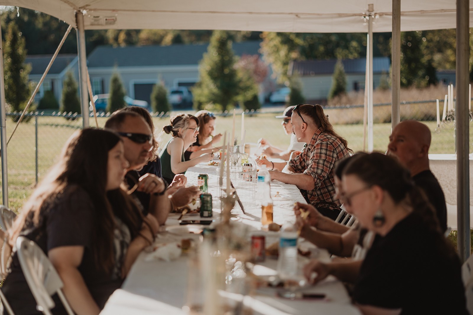 Guests eating and talking down one long banquet table under the tent, low gold sunlight coming in sideways under the canopy