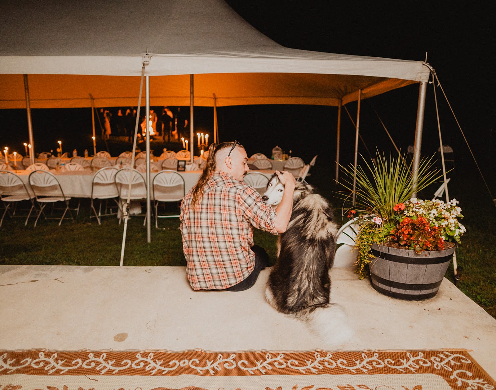 A guest kneeling on the patio at night with both arms around the malamute, the candlelit tent glowing behind them