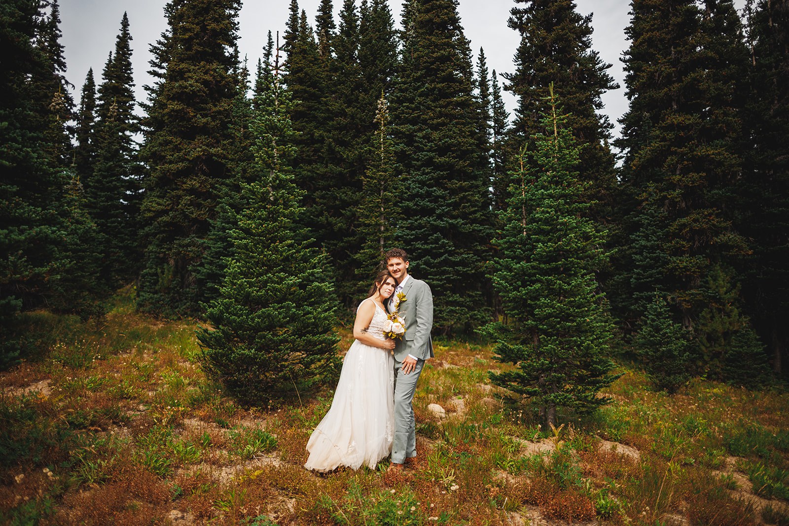 Michaela and AJ standing small in a subalpine meadow, ringed on every side by tall dark firs under a pale sky