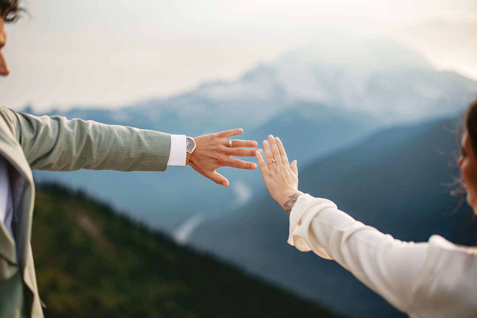 Michaela and AJ reaching their ringed hands toward each other on the summit at golden hour, the snow dome of Mount Rainier filling the gap between their hands