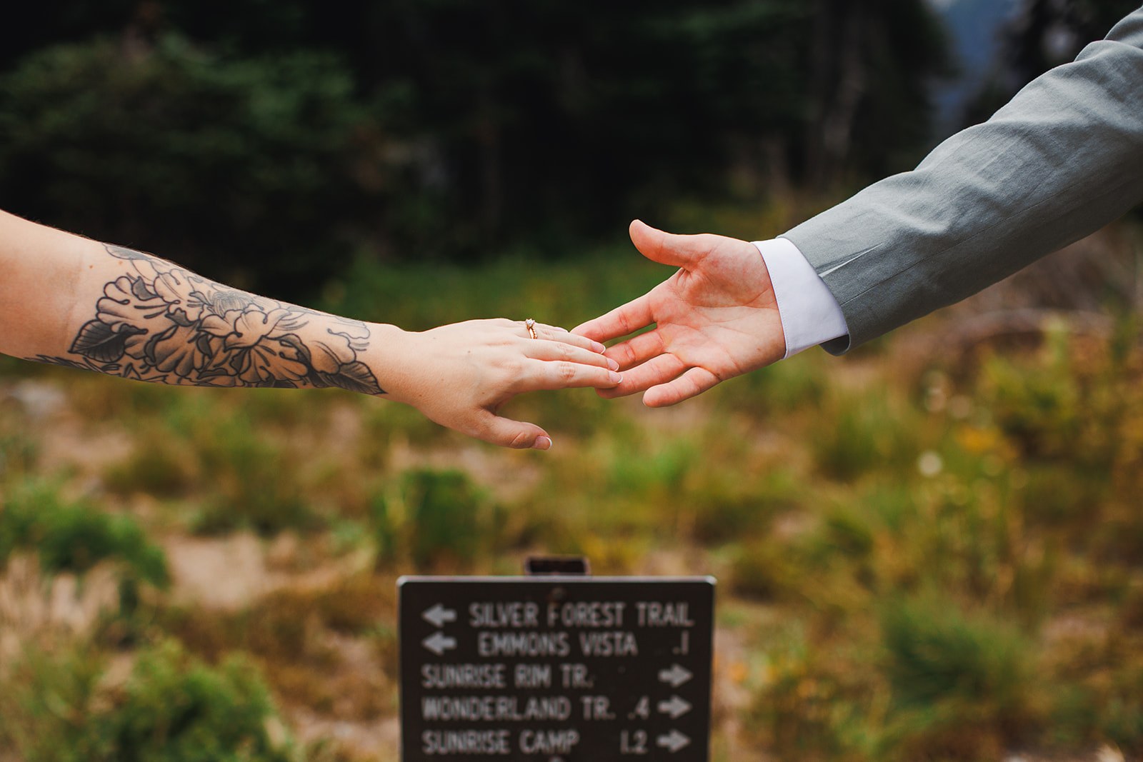 Michaela and AJ reaching their newly ringed hands toward each other above a trail junction sign reading Silver Forest Trail, Emmons Vista, Sunrise Rim Trail, Wonderland Trail, Sunrise Camp