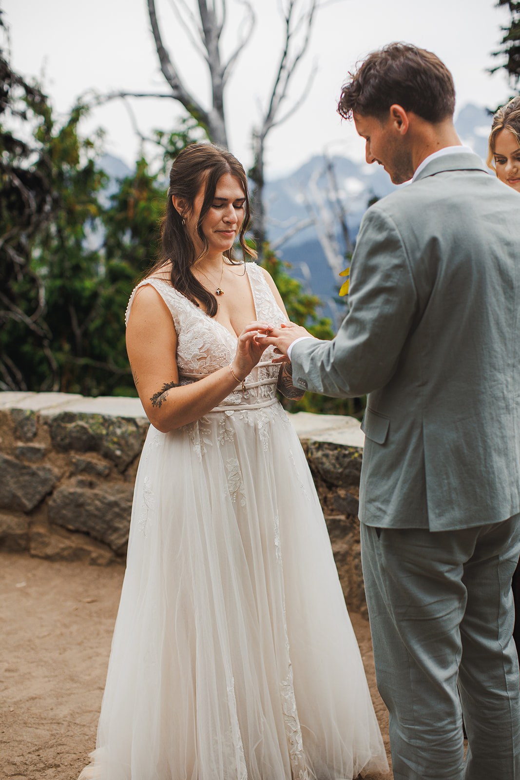 Michaela sliding a ring onto AJ's finger during the ceremony, her lace dress against his sage linen suit, the stone overlook wall behind them