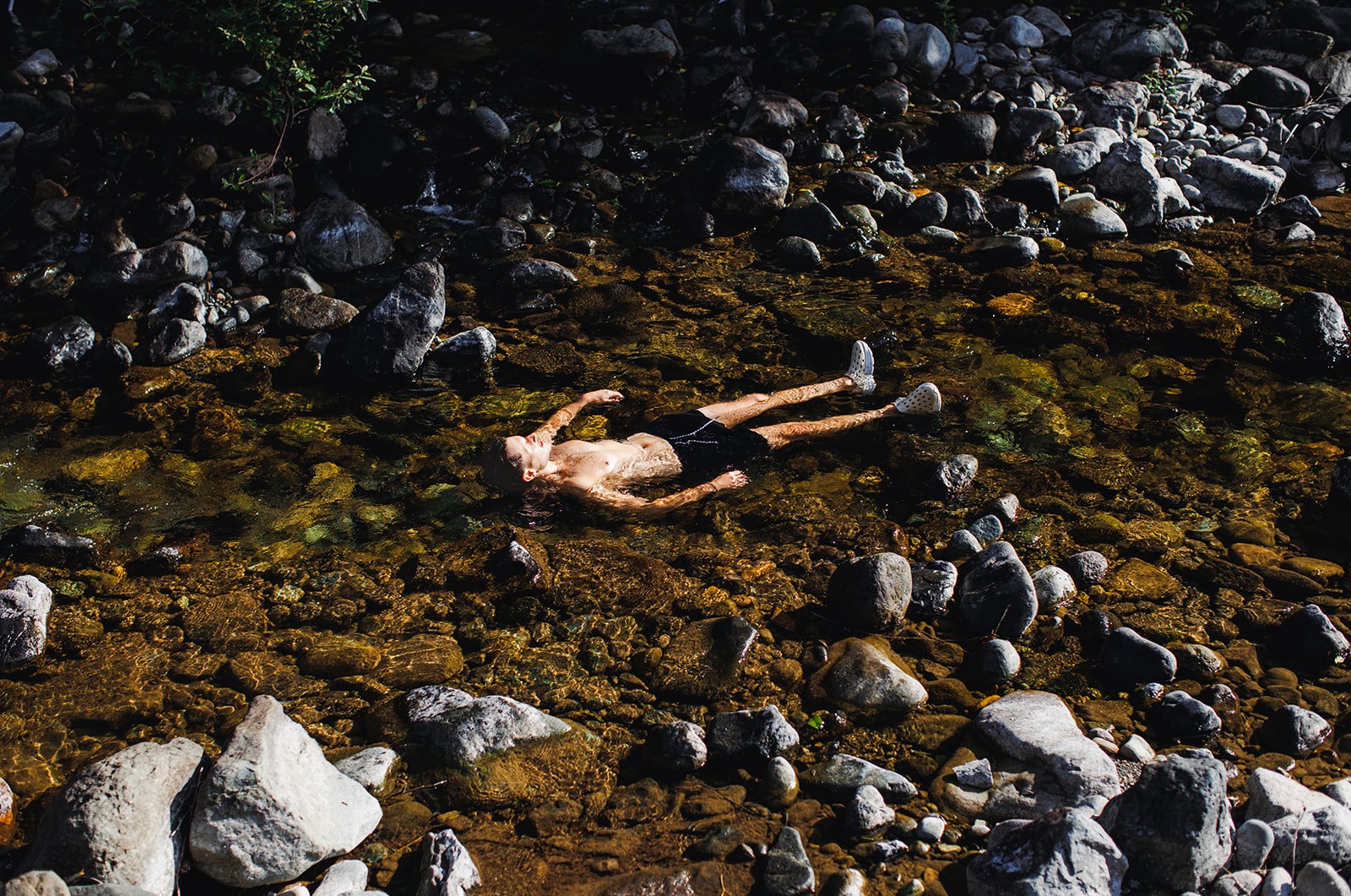 AJ floating on his back in a few inches of clear water on the morning of his wedding, seen from above, surrounded by the pale river stones of the shallows