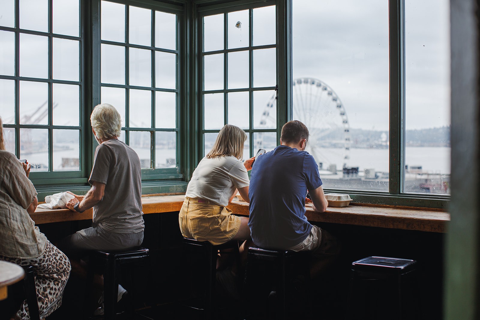 The Seattle Great Wheel seen through the tall green windows of the Waterfront Viewpoint, people at the counter looking out over Elliott Bay