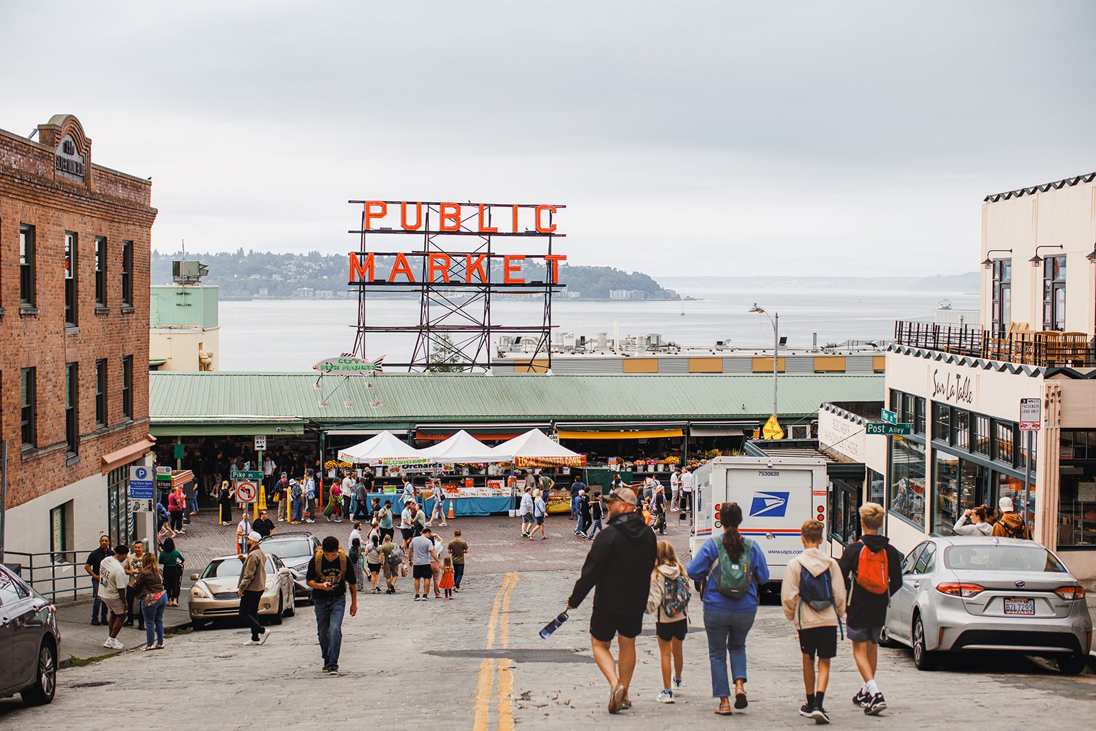 The red Public Market Center neon sign above Pike Place Market in Seattle, the cobbled street full of people below it and Elliott Bay beyond