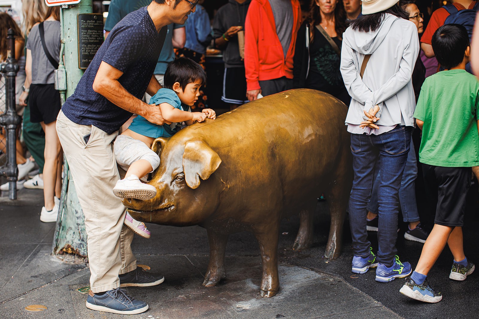 A father lifting a toddler onto Rachel the bronze piggy bank at the entrance to Pike Place Market while other children wait their turn