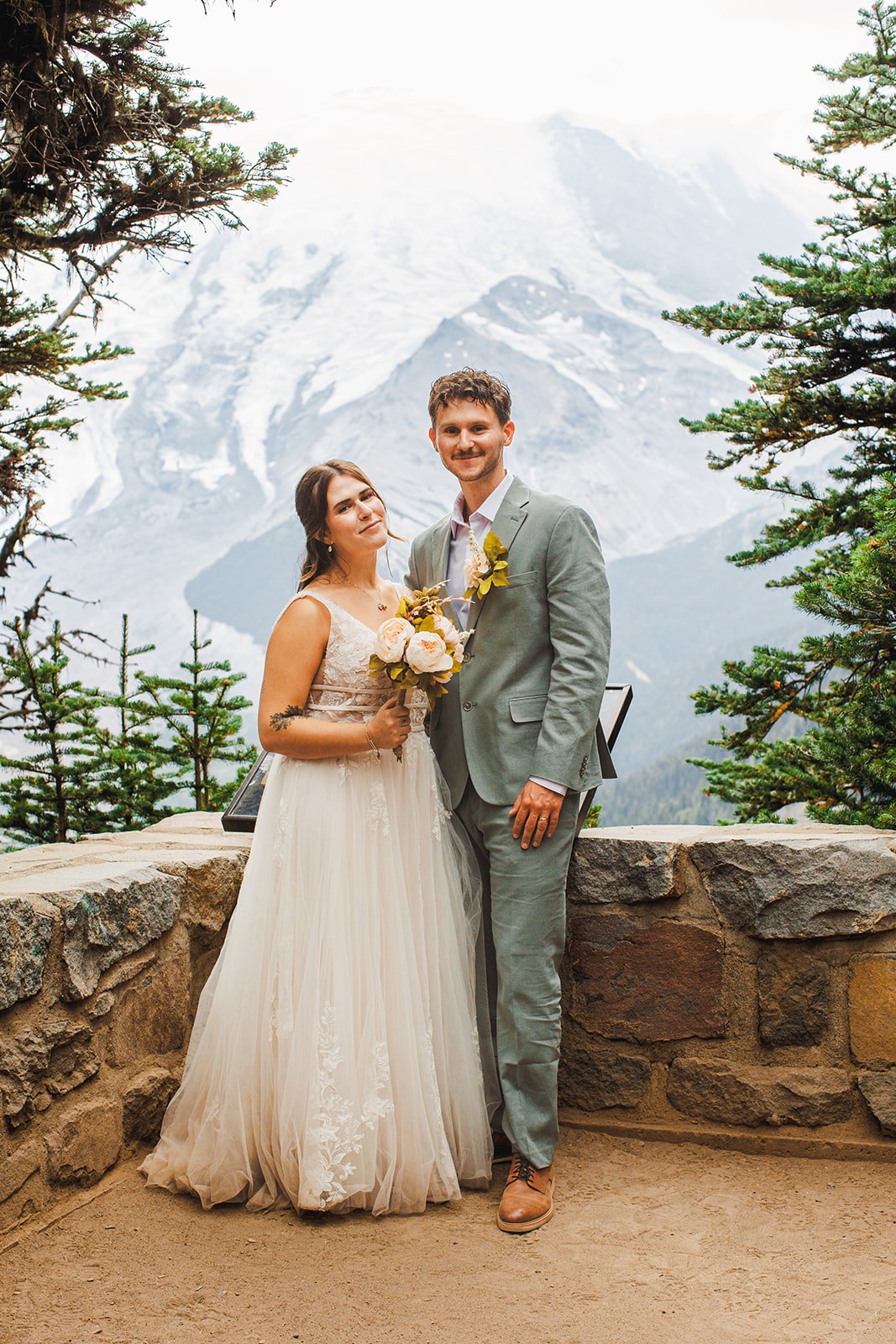 Michaela and AJ at the overlook wall just after the ceremony, her bouquet at her waist, the snowfields of Mount Rainier filling the sky behind them
