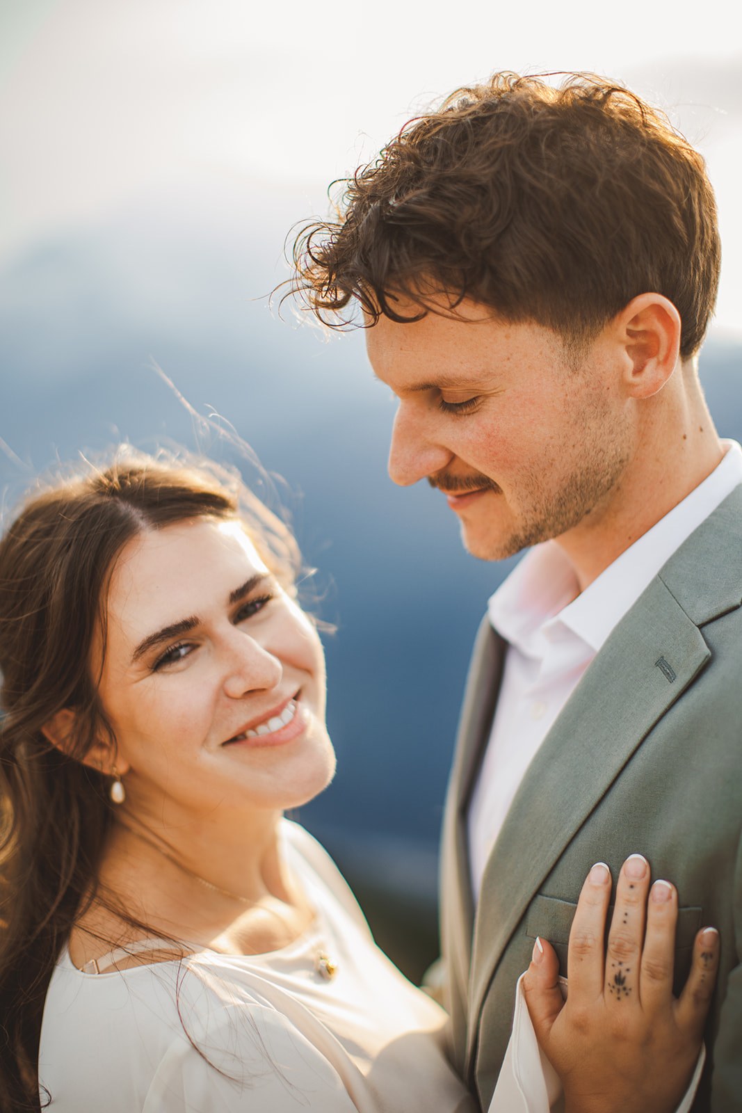A close portrait of Michaela smiling with her hand flat on AJ's chest at the summit, both of them lit by low hazy evening sun
