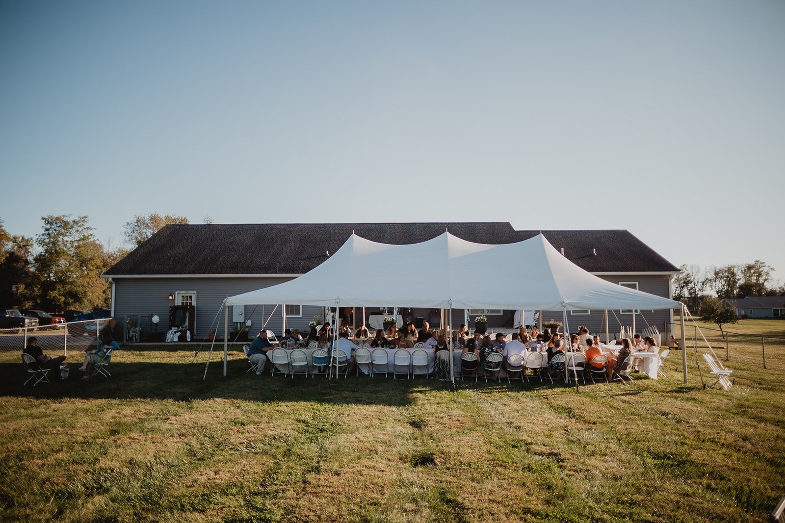 The white pole tent on the back lawn at golden hour, both long tables full of seated guests, the house and open field behind