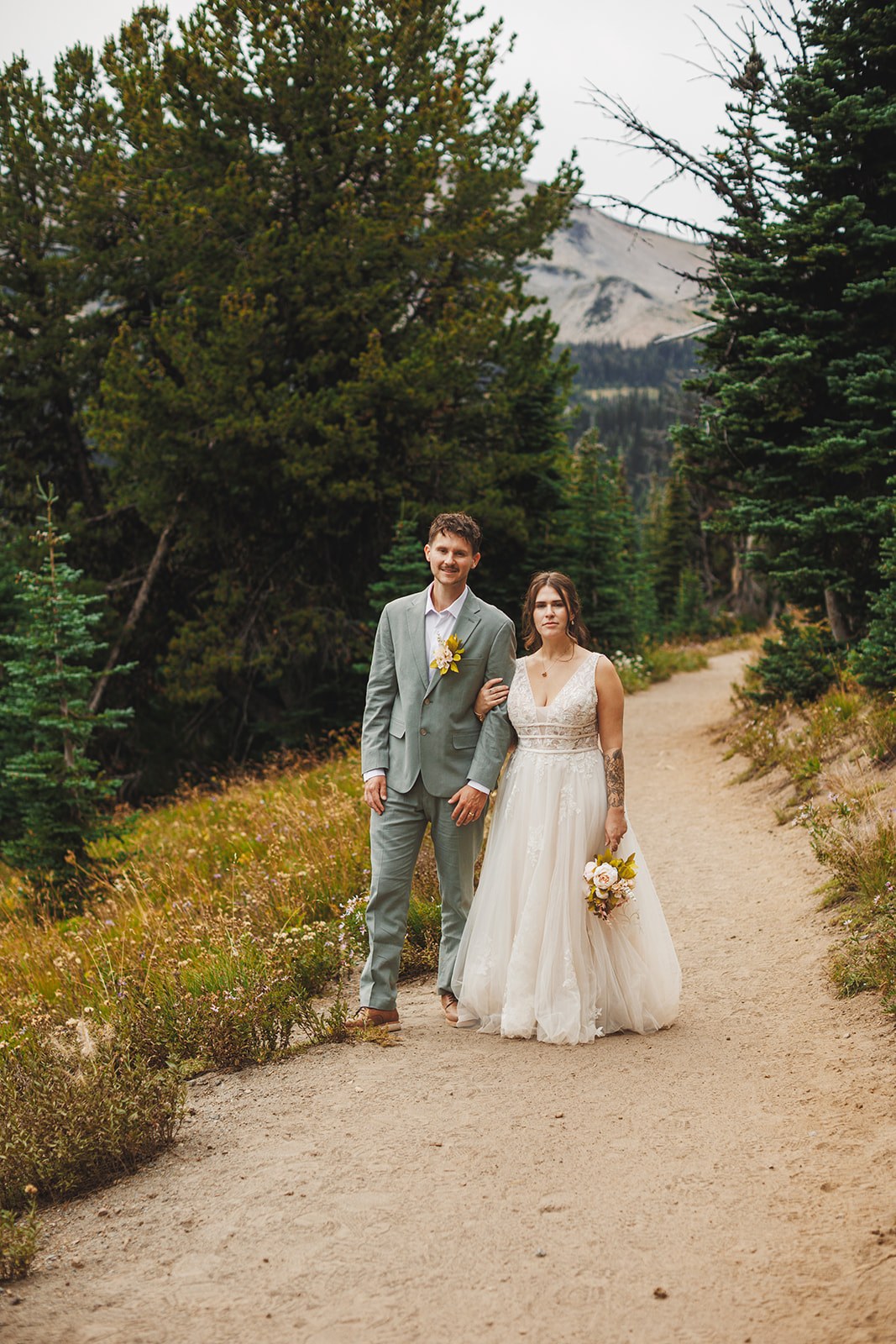 Michaela and AJ standing arm in arm on the gravel trail with meadow on both sides and a bare ridge behind them