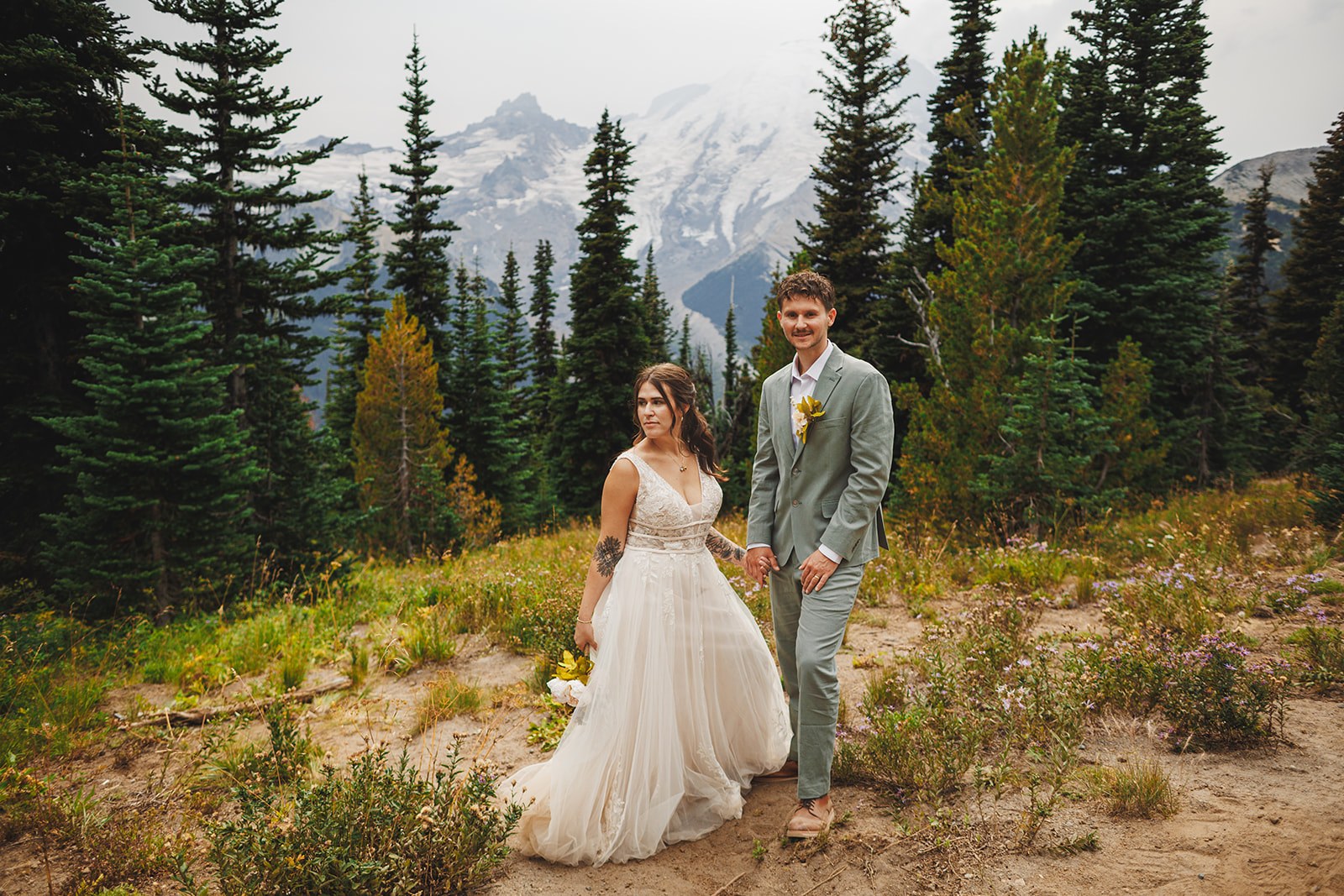 Michaela and AJ standing together on the Silver Forest Trail in subalpine meadow, dark evergreens on both sides and the snow and rock of Mount Rainier rising behind