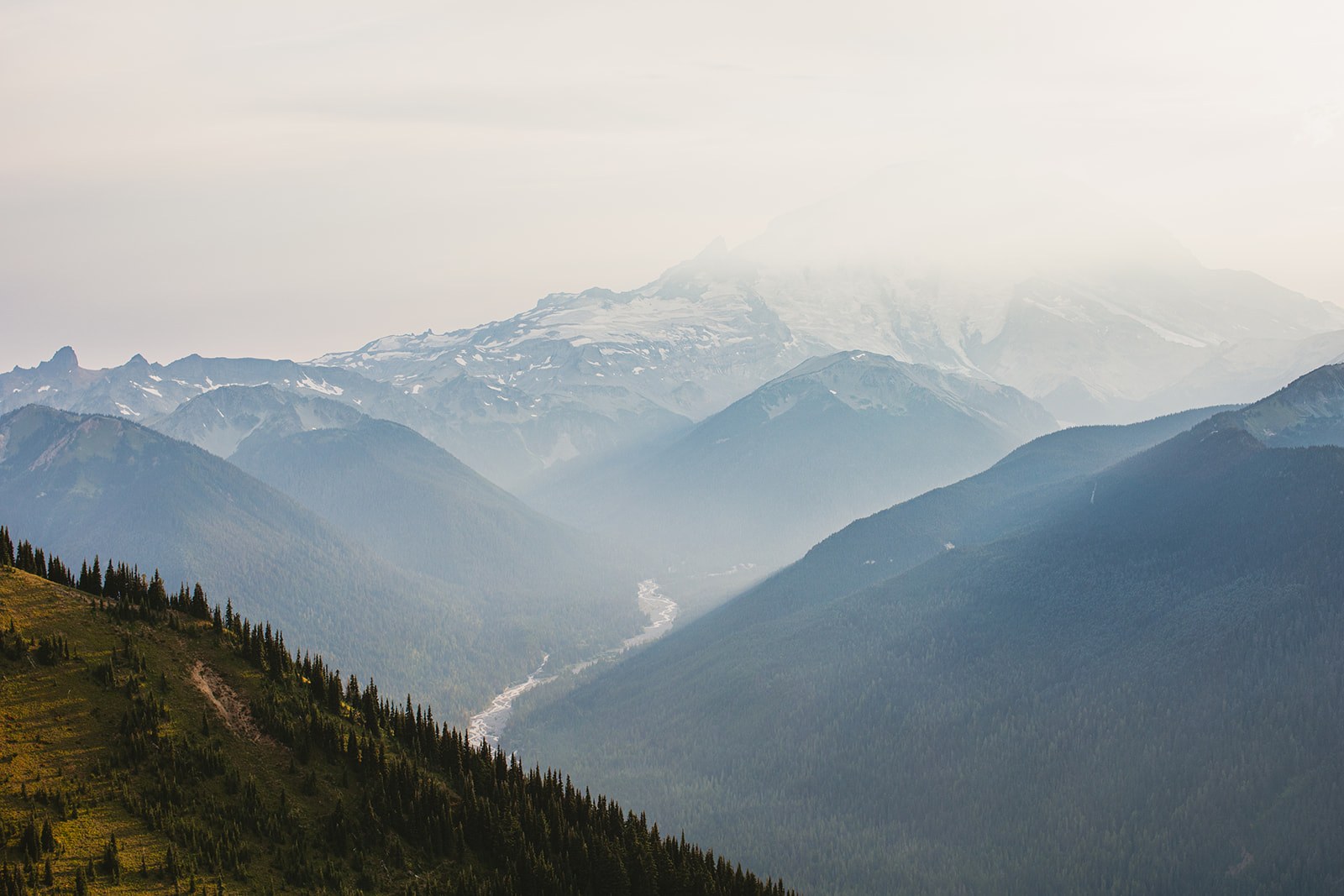 Hazy blue ridges falling away from the summit with a river valley far below and Mount Rainier's snow dome behind