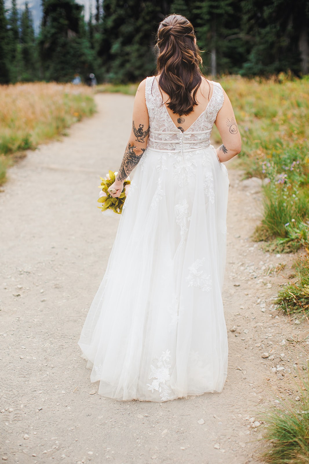 Michaela walking away up the gravel trail at Sunrise, seen from behind, the low open back of her lace dress and her bouquet at her side