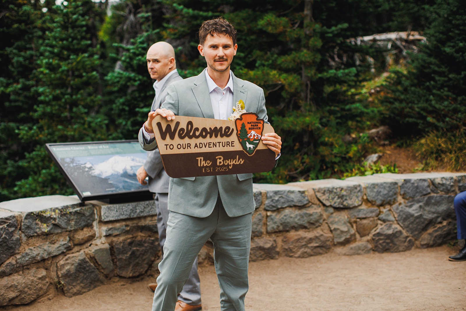 AJ in his sage linen suit carrying a wooden sign shaped like a National Park Service arrowhead that reads Welcome to our adventure, The Boyds, est. 2025