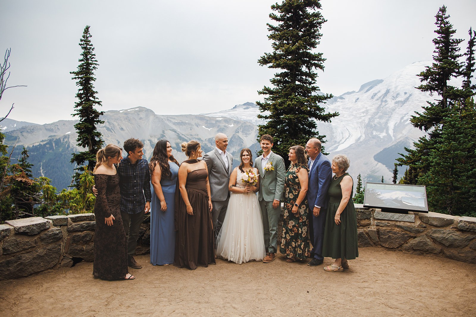 The entire guest list, ten people, lined up along the stone overlook wall at Emmons Vista with Mount Rainier behind them