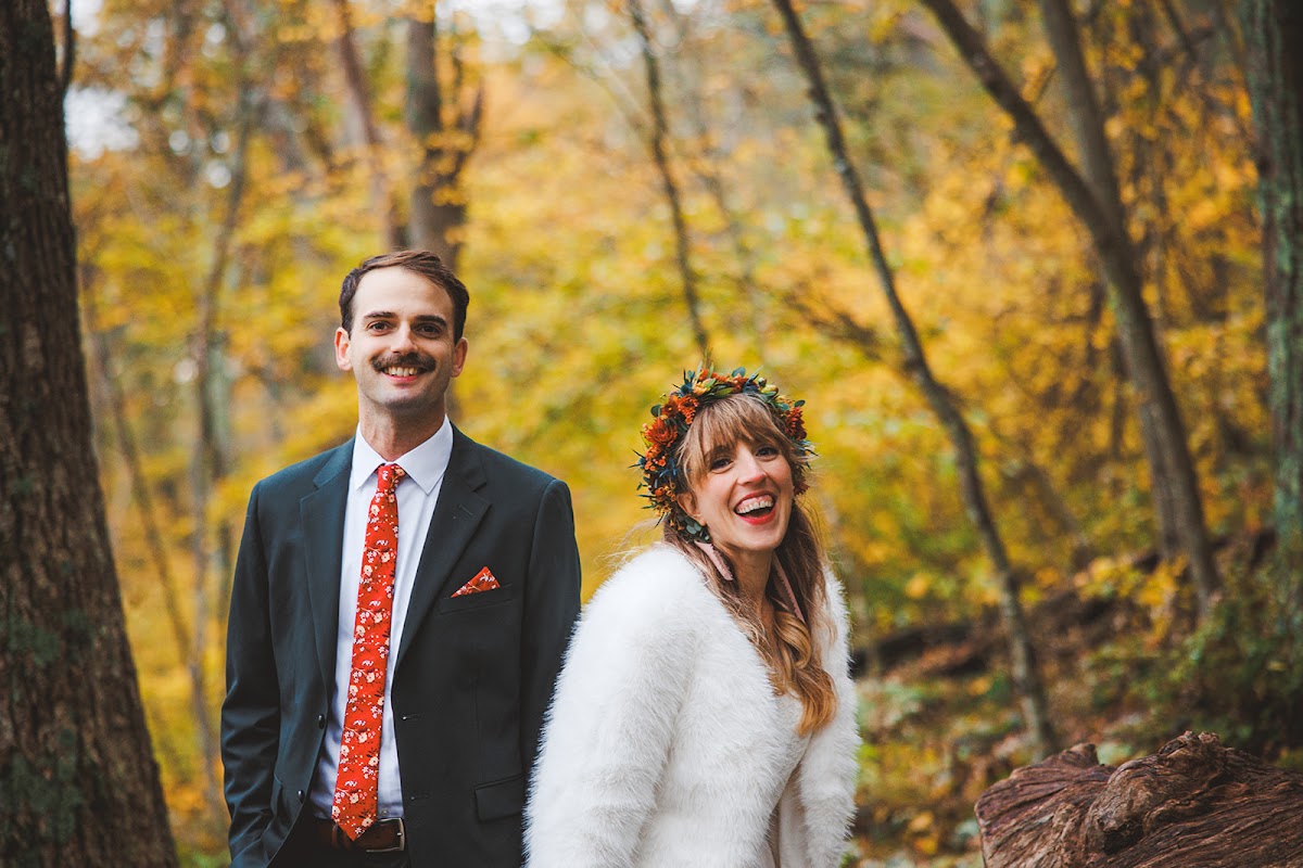 Sara and Ryan standing side by side in the golden autumn woods, her flower crown and fur jacket against his dark green suit and orange floral tie