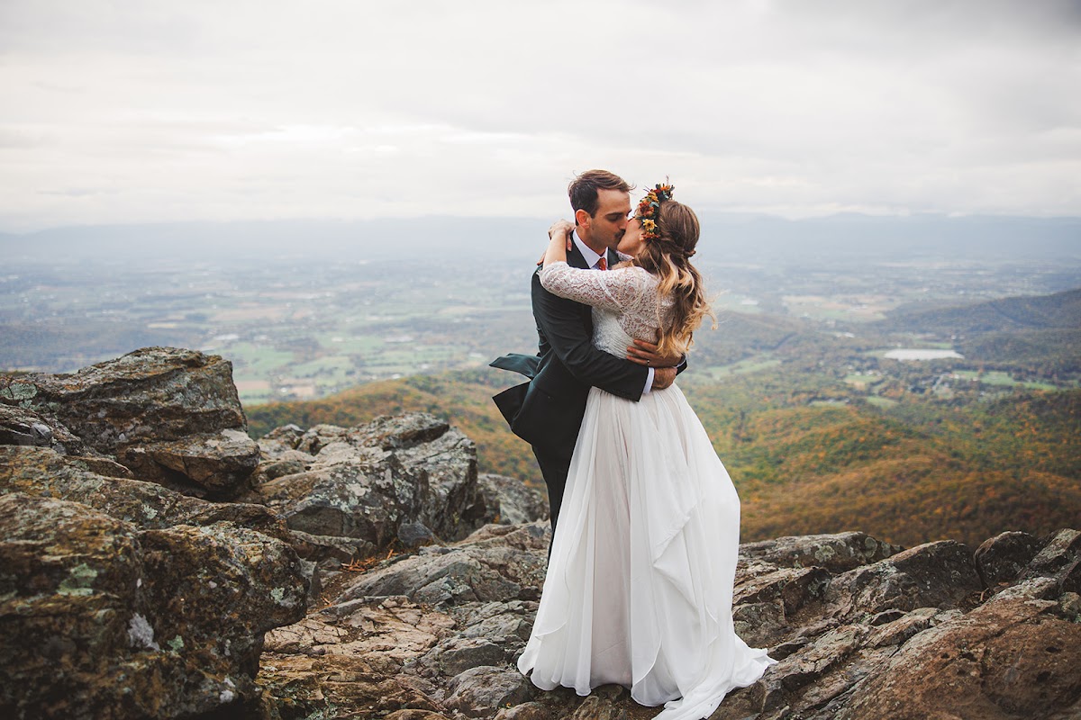 Sara and Ryan kissing on the rock ledge, her arms around his neck, the valley floor and distant ridgelines behind them