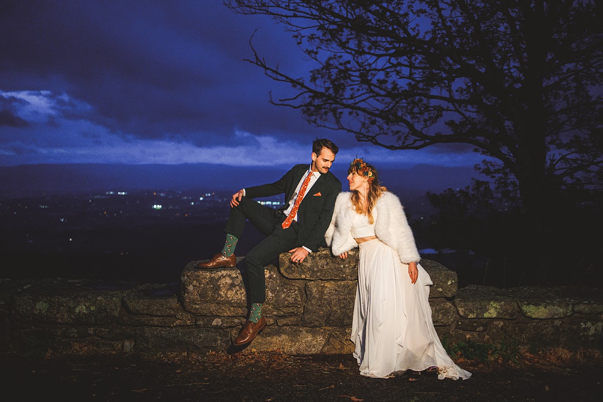 Sara and Ryan sitting relaxed on a low stone wall at dusk, leaning toward each other, faint valley lights and a bare tree behind them