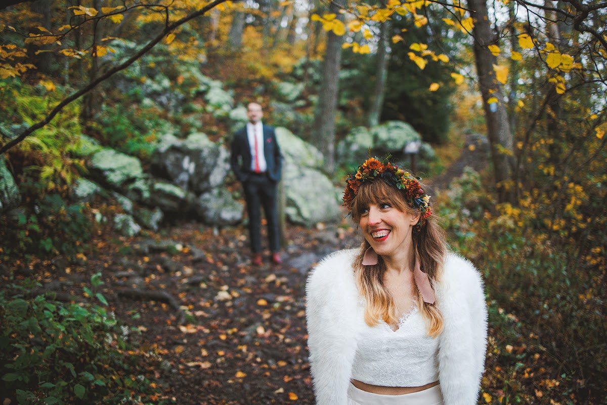 Sara laughing in her fur jacket and flower crown on the trail, Ryan soft-focus on the rocks behind her with his hands in his pockets