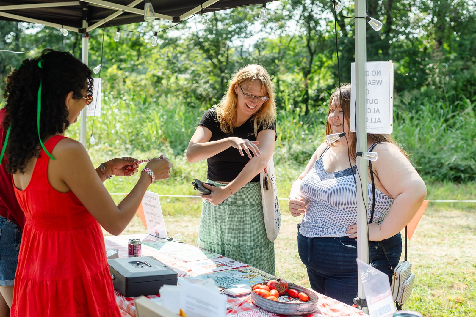 A volunteer in a red dress fastening wristbands for two arriving guests at the Tomato Jam check-in table