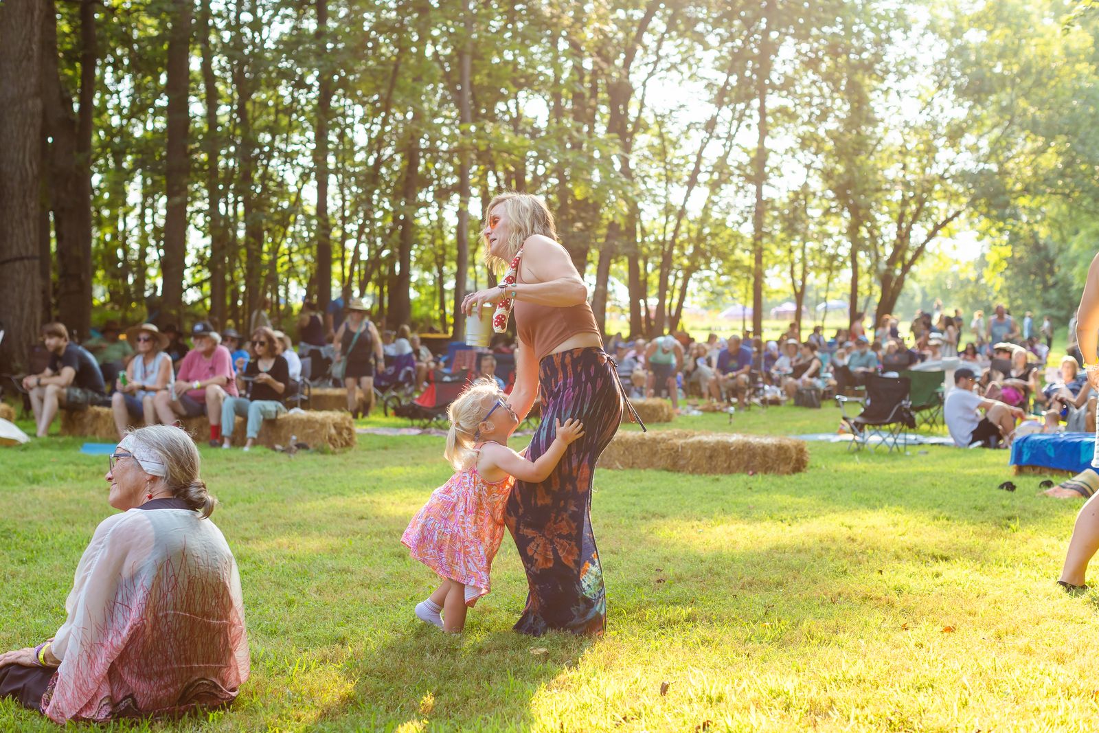 Backlit by low golden sun, a woman in a patterned skirt dances with a toddler in a pink dress on the open grass in front of the crowd
