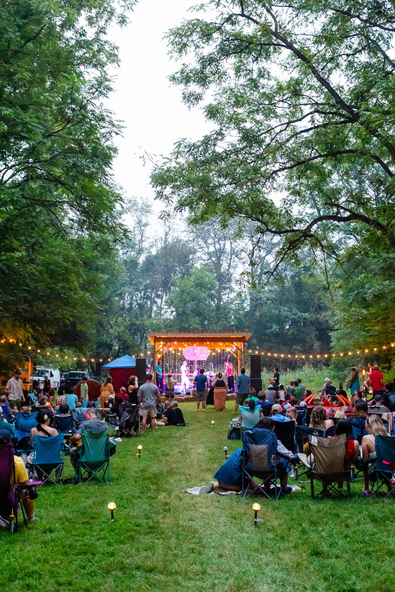 Dusk view down the tree-lined lawn at Sistermoon Farm toward the lit Tomato Jam stage, the crowd seated along a grassy aisle