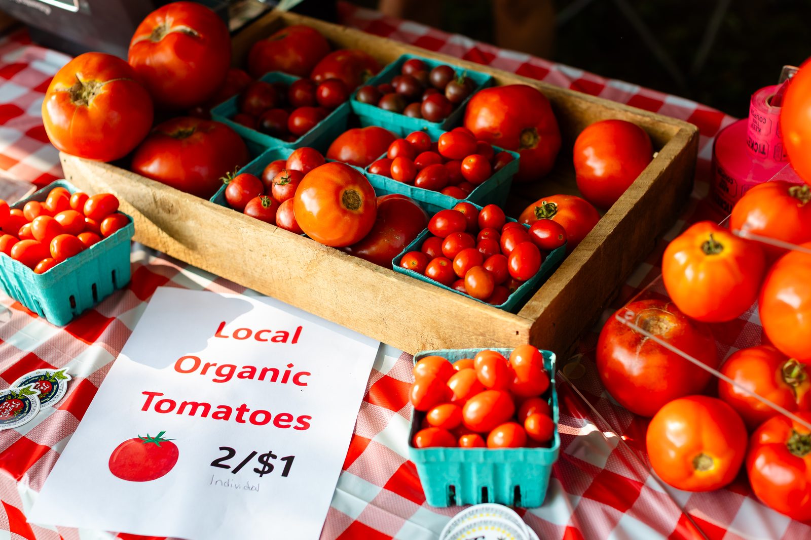 Crates of ripe local tomatoes on a gingham tablecloth behind a sign reading Local Organic Tomatoes, two for a dollar