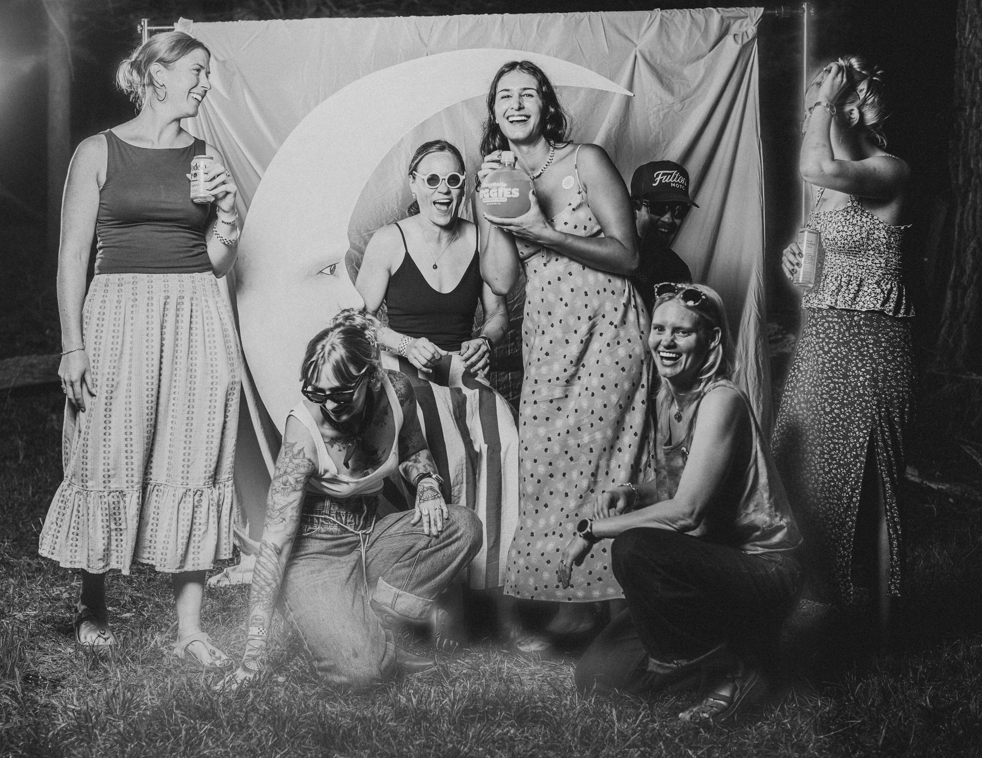 Seven friends laughing around the crescent moon at the Tomato Jam Moon Booth, one drinking from a disco ball cup, in black and white