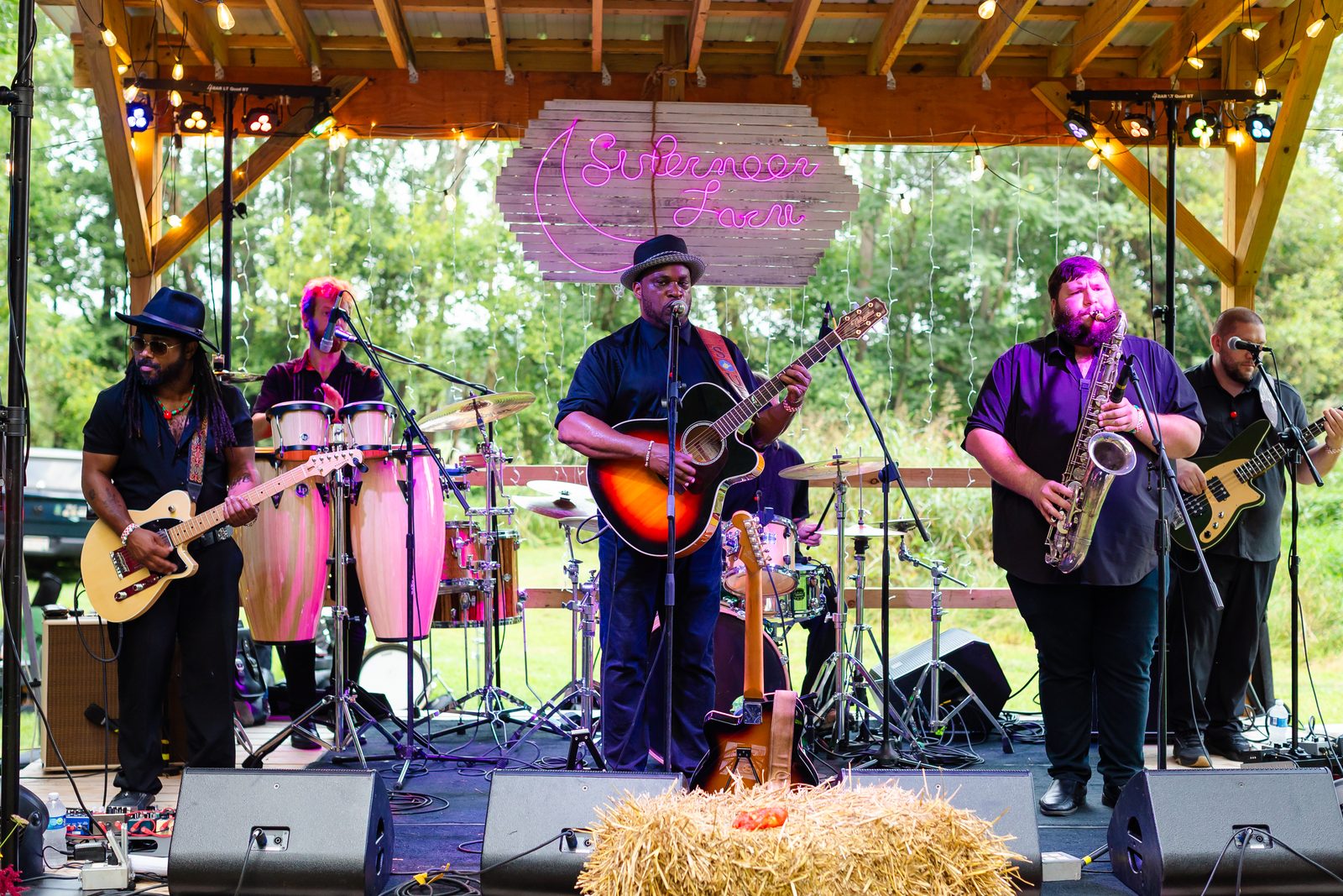 A full band with congas and saxophone playing under the string-lit pavilion at night, hay bales at the stage lip