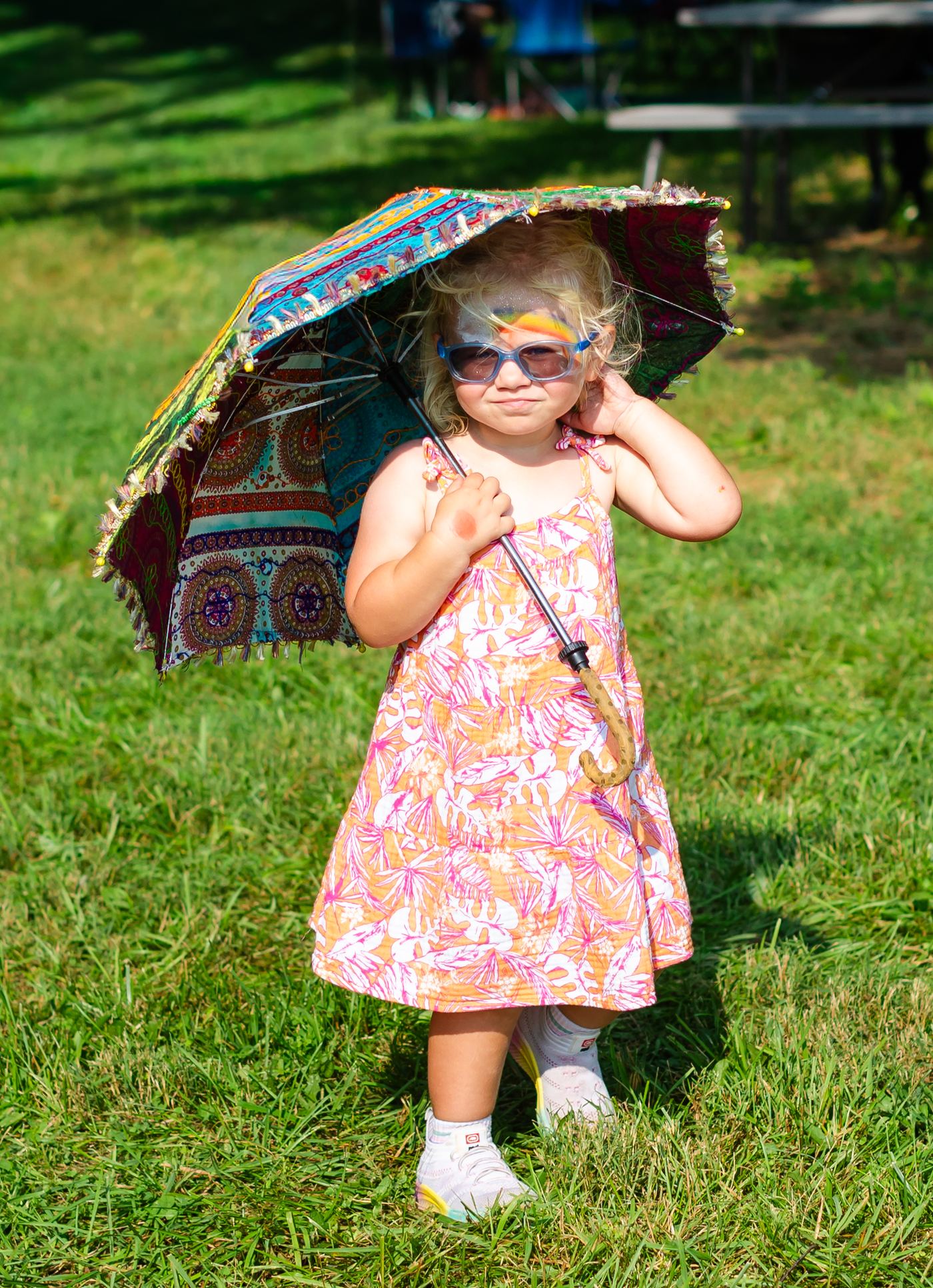 A toddler in a sundress and yellow sunglasses holding an embroidered parasol on the grass at Tomato Jam