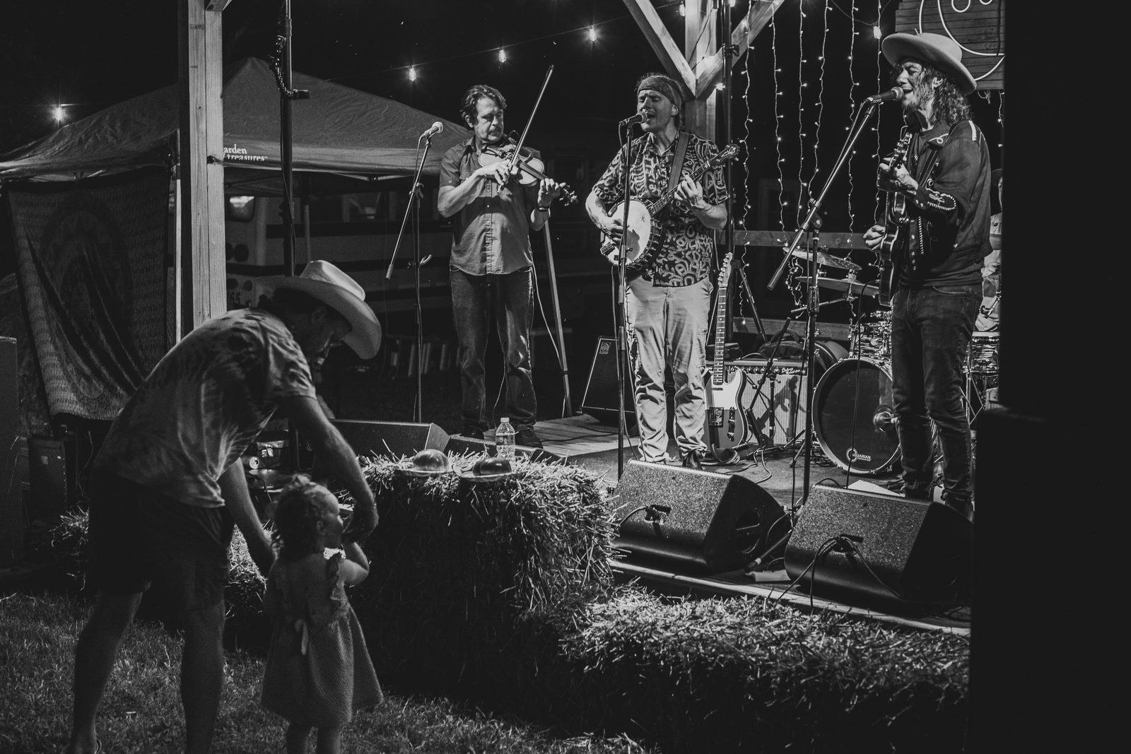 Black and white frame of the string band playing while a parent in a cowboy hat steadies a toddler standing on a hay bale
