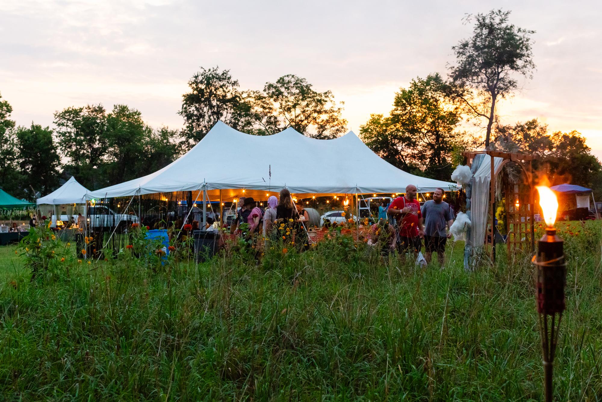 The white sailcloth vendor tent glowing at sunset across a meadow of tall grass and wildflowers at Sistermoon Farm, a tiki torch burning at the edge