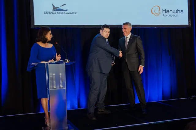 Award recipient shakes hands with a presenter on stage beside a host at a glass lectern under blue lighting.