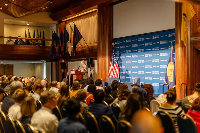 Rows of attendees fill a wood-paneled hall facing a blue press backdrop.