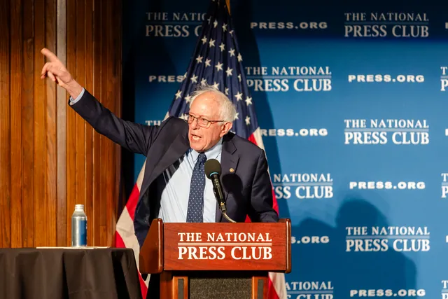 Bernie Sanders at the National Press Club podium with his arm raised, the flag and press club backdrop behind him.