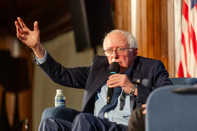 Bernie Sanders seated on stage, mid-gesture with a microphone, in front of an American flag.