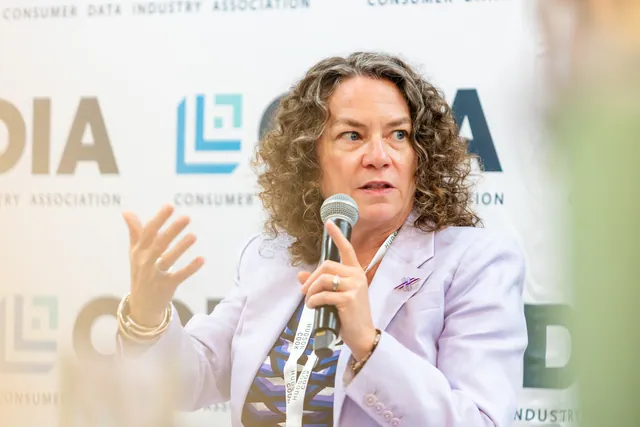 Speaker in a lavender blazer gesturing with a microphone against the branded backdrop.