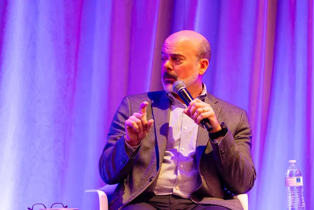 Panelist gesturing with a microphone against purple stage lighting.
