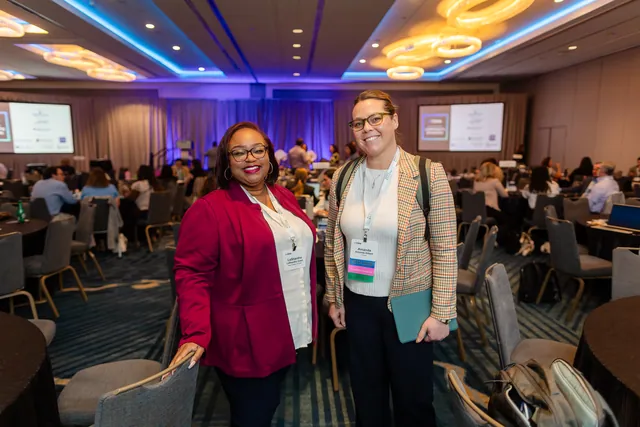 Two attendees smiling at the camera in a crowded ballroom.