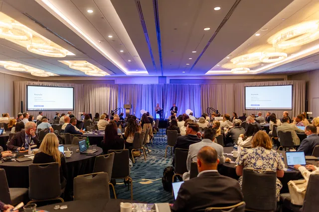 Wide ballroom view of a full crowd facing a lit stage with screens.