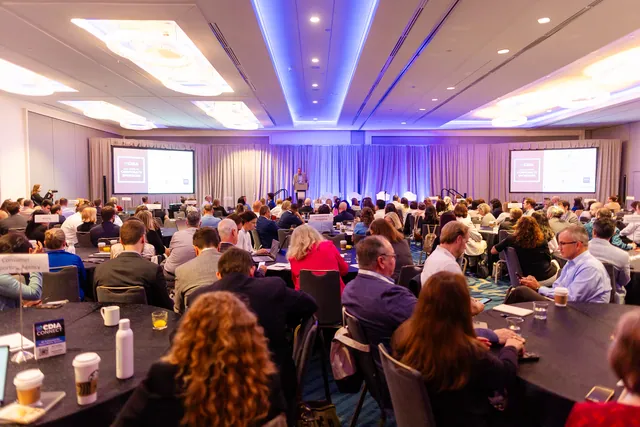Packed general session audience facing the stage in a large ballroom with blue ceiling lighting.