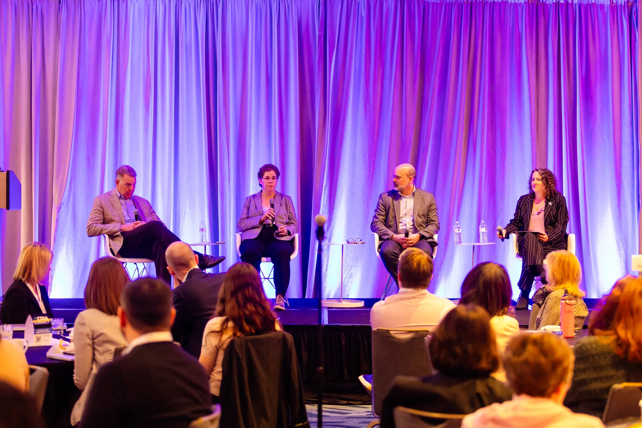 Four panelists seated on stage in front of glowing purple curtains as the audience looks on.