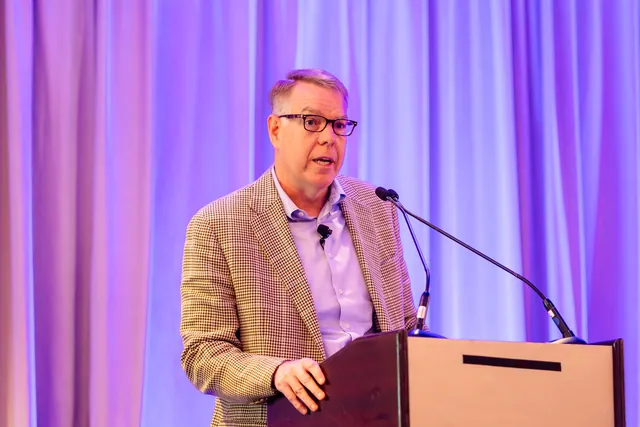 Speaker in glasses and a tweed blazer talking at a podium against purple curtains.